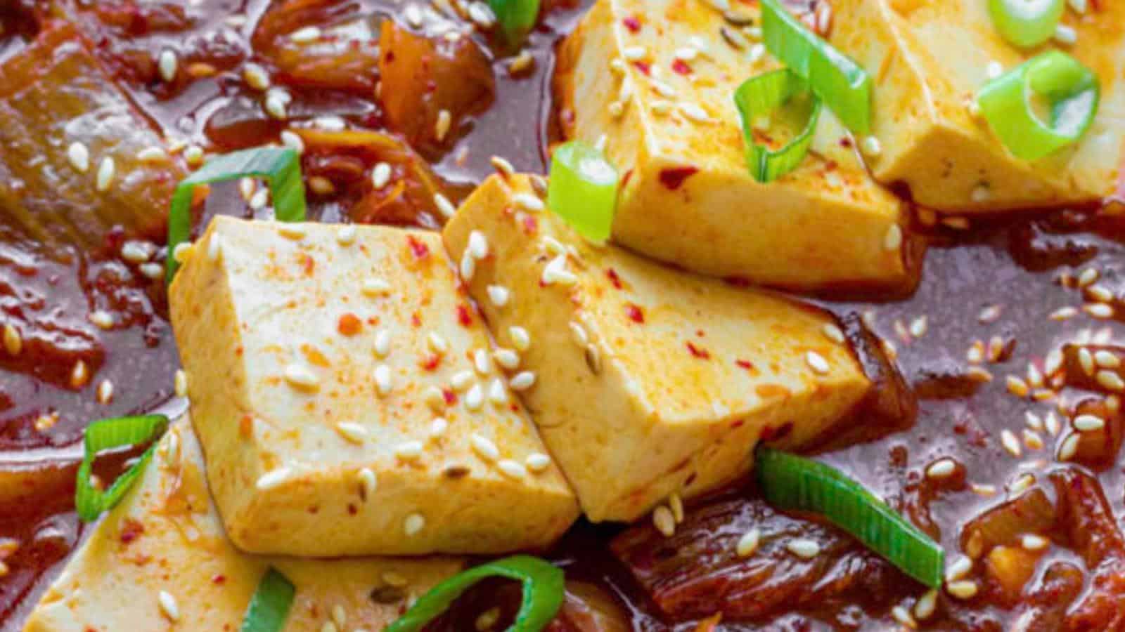 Close-up of a spicy tofu dish garnished with sesame seeds and sliced green onions. The tofu is bathed in a rich, red sauce, with visible chunks of onions. The vibrant colors and garnishes highlight the dish's appetizing and flavorful presentation.
