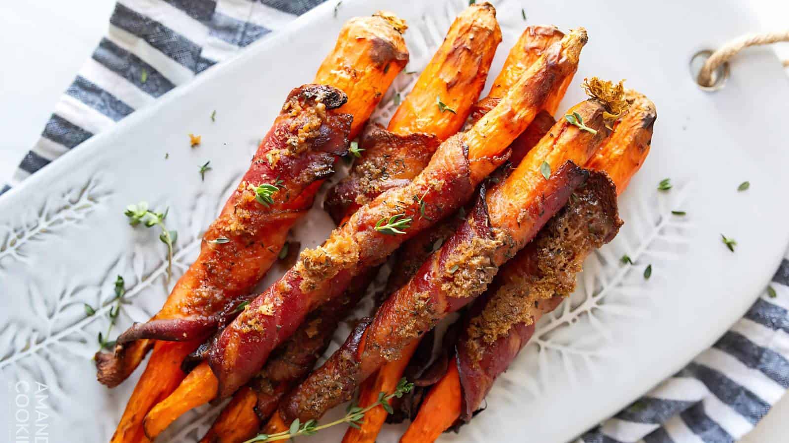 A plate of roasted carrots wrapped in bacon, garnished with herbs, on a decorative white dish with a striped cloth underneath.