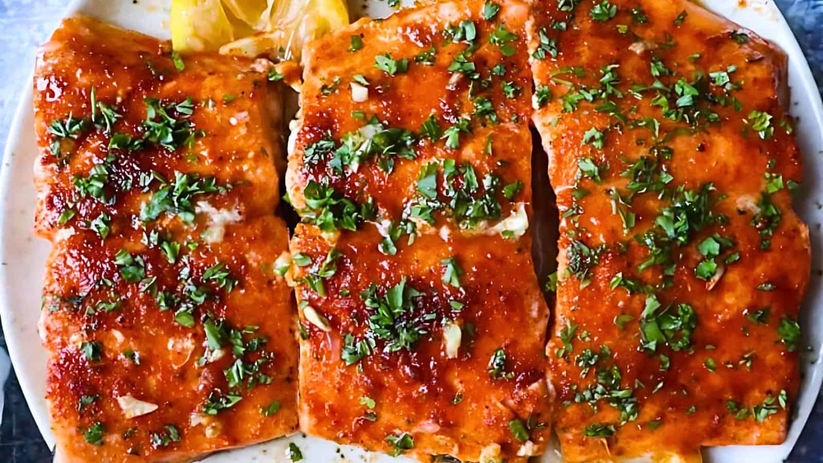 The image shows three pieces of grilled salmon, covered in sauce, garnished with chopped herbs, and accompanied by lemon wedges on a white plate.