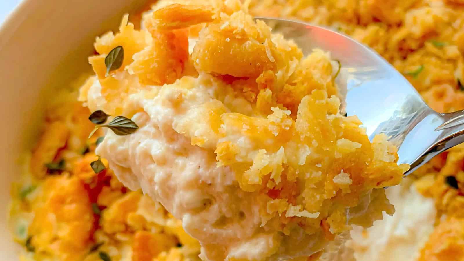 A spoonful of a cheesy, creamy dish with a crispy golden topping, garnished with small fresh herbs. In the background, the rest of the dish is visible in a bowl.