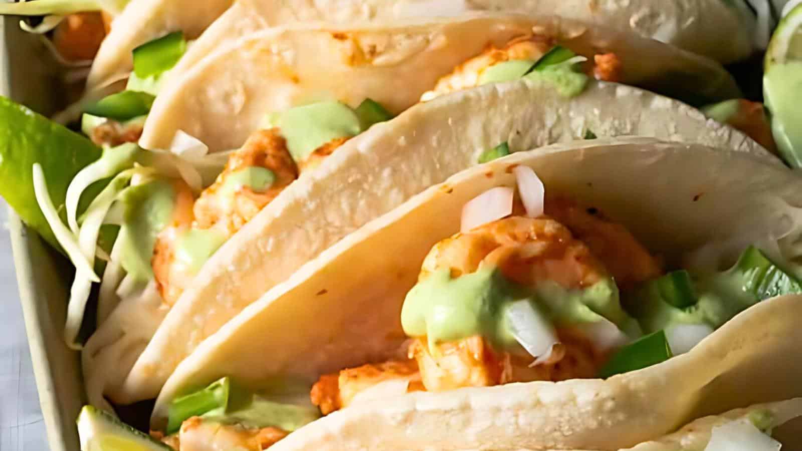 Close-up of a tray filled with shrimp tacos topped with white onions, chopped green peppers, and creamy avocado sauce. The tacos are served in folded, lightly toasted tortillas, with a wedge of lime partially visible on the side.