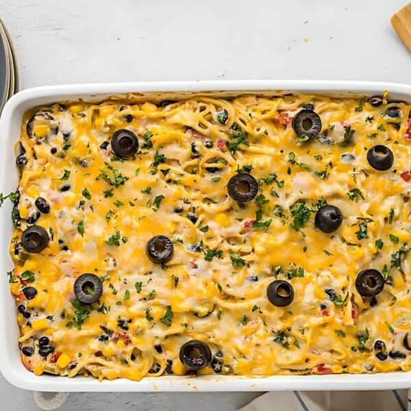 A baked casserole dish filled with cheesy pasta, black olives, and herbs. Nearby are plates, forks, a wooden spatula, and a towel on a light-colored surface.