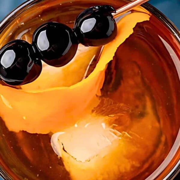 A large ice cube, an orange peel garnish, and three black cherries skewered on a cocktail pick.