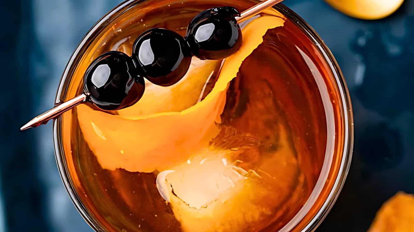 A large ice cube, an orange peel garnish, and three black cherries skewered on a cocktail pick.