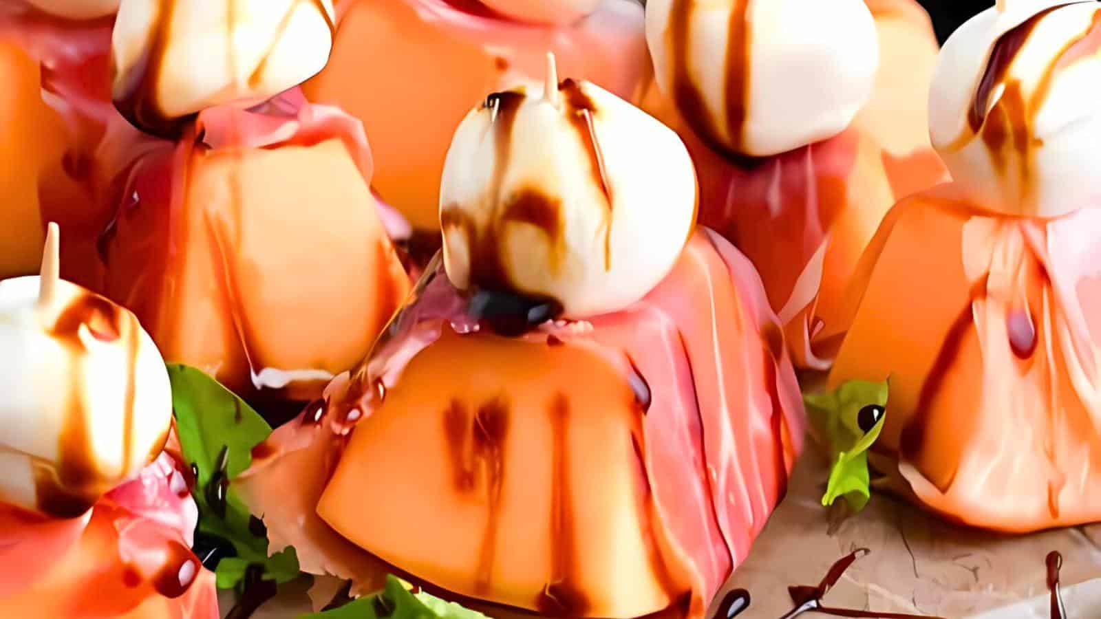 A display of melon topped with small white dollops, drizzled with a dark liquid, arranged on a plate garnished with green leaves.
