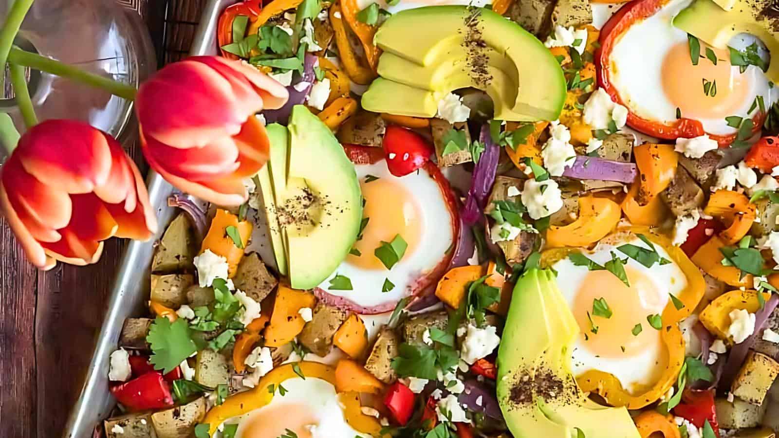 The image shows a colorful dish with baked eggs, avocado slices, chopped vegetables, and herbs on a tray. Beside it is a vase containing three red tulips.