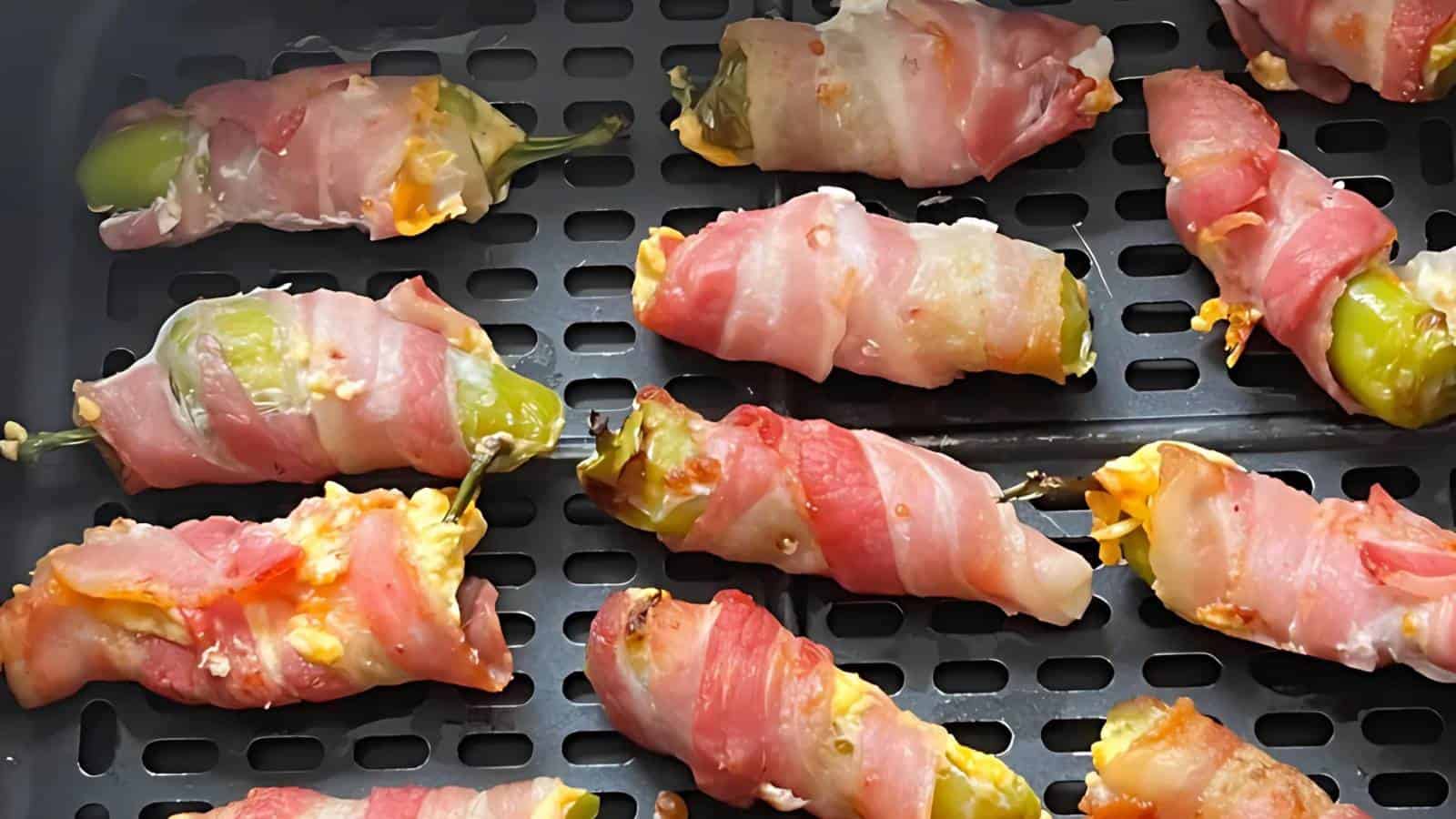 Jalapeño peppers stuffed with cheese and wrapped in bacon are arranged on an air fryer tray.