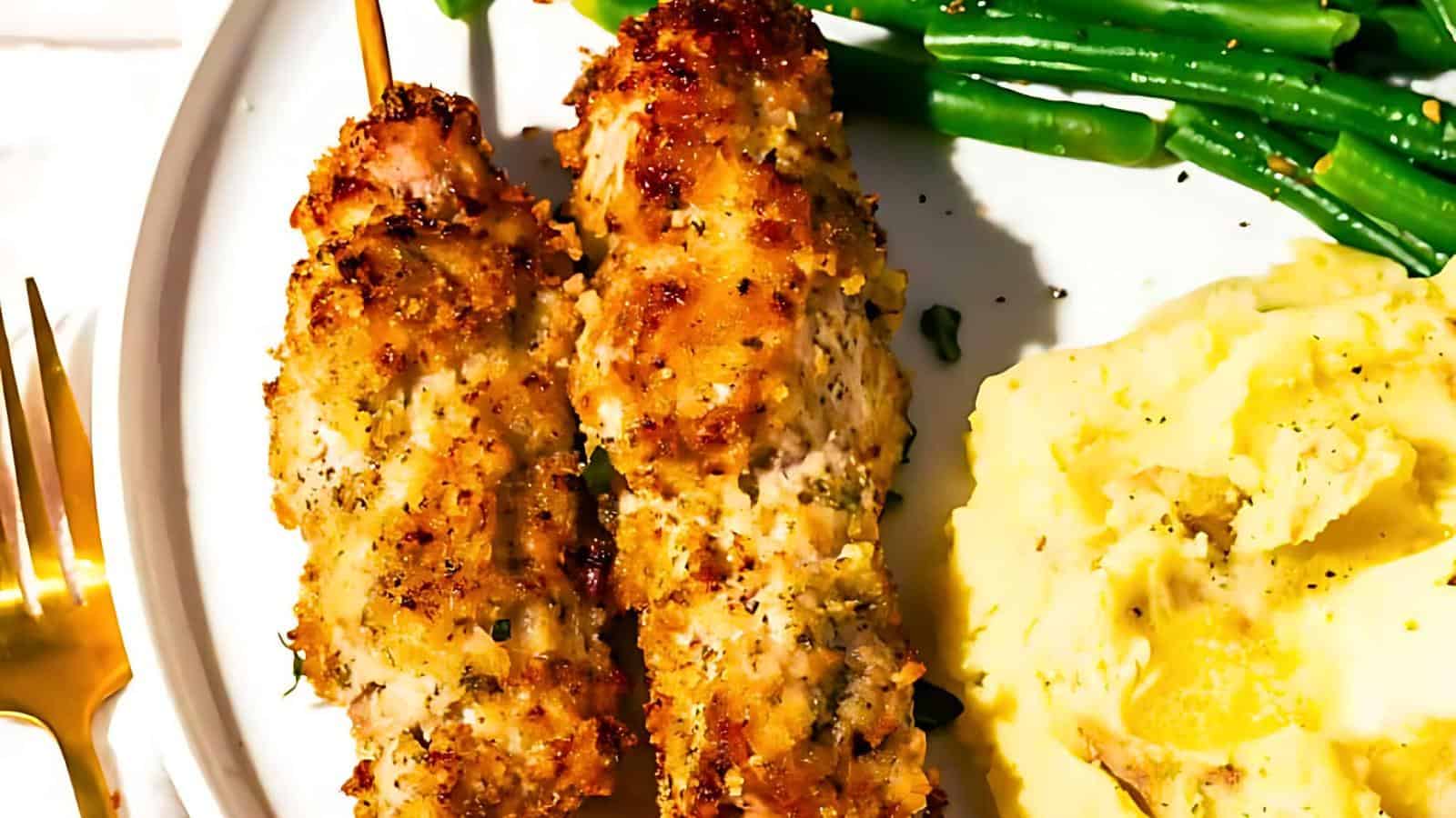 Two skewered chicken pieces coated in breadcrumbs, served with mashed potatoes and green beans on a white plate with a gold fork.