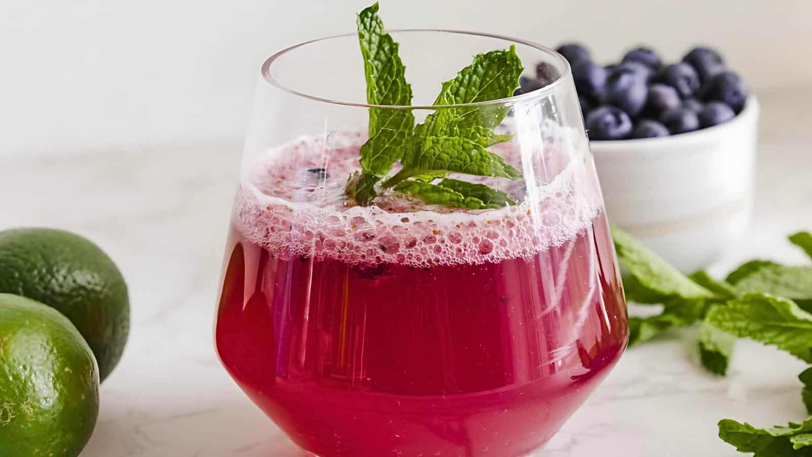 A glass of red sparkling drink garnished with mint leaves, placed on a white surface next to whole limes, scattered mint leaves, and a bowl of blueberries in the background.
