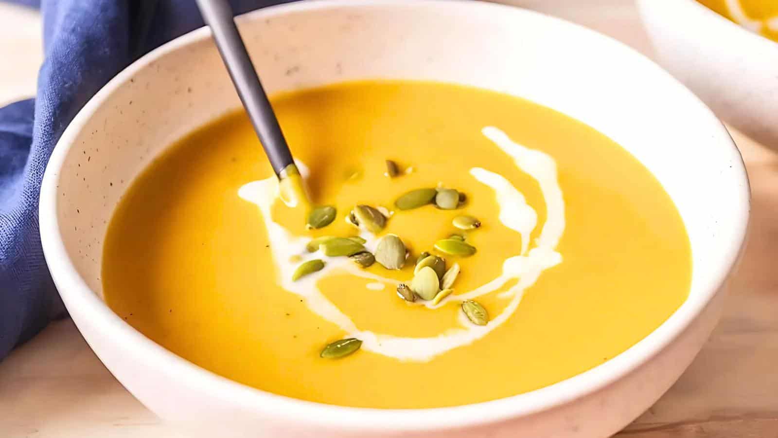 A bowl of creamy soup topped with a swirl of white cream and sprinkled with pumpkin seeds, with a spoon resting in the bowl.