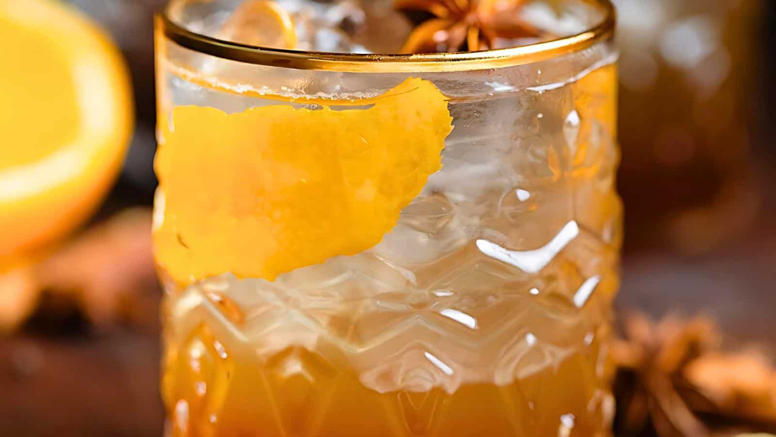 A textured glass filled with a cocktail, featuring a large orange peel garnish and ice cubes. The background includes a lemon and star anise, slightly blurred.