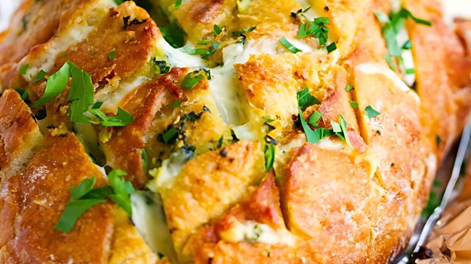 A close-up of a loaf of pull-apart bread with melted cheese and sprinkled with chopped herbs, displaying a golden-brown crust and appetizing texture.