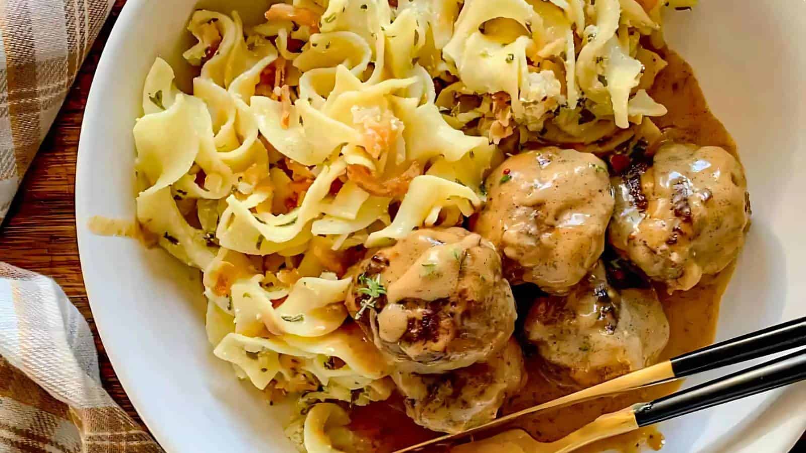 A bowl of creamy Swedish meatballs with brown gravy sauce, served with egg noodles and garnished with herbs. Chopsticks rest on the bowl's edge. A brown plaid cloth is beside it.