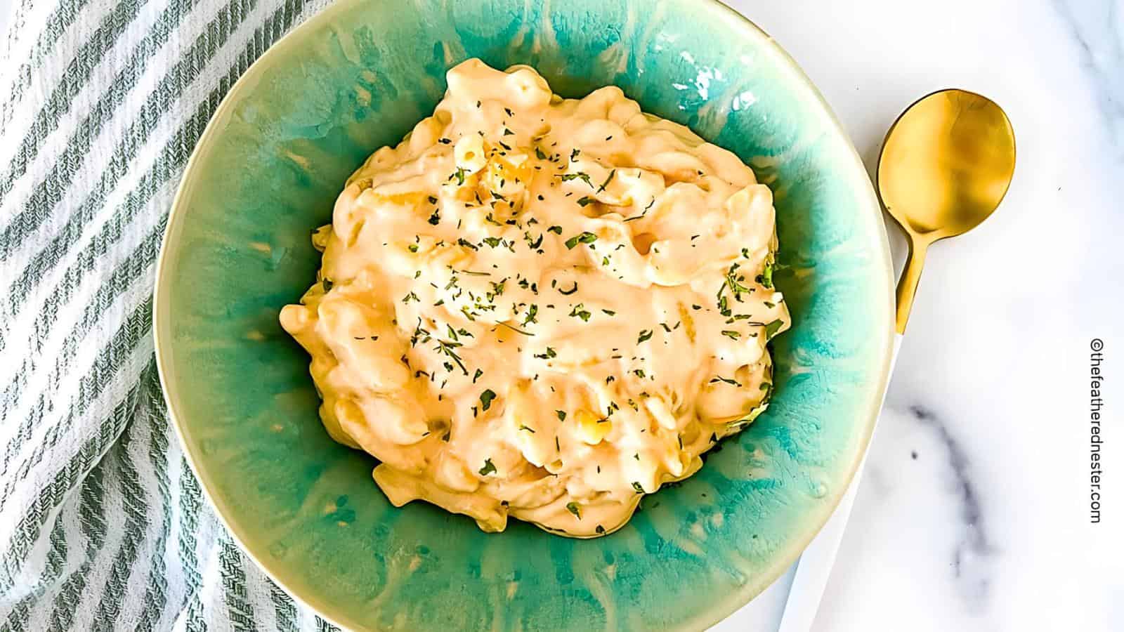 A bowl of creamy mac and cheese garnished with herbs on a green plate. A gold spoon on a white surface to the right. A striped cloth partially visible to the left.