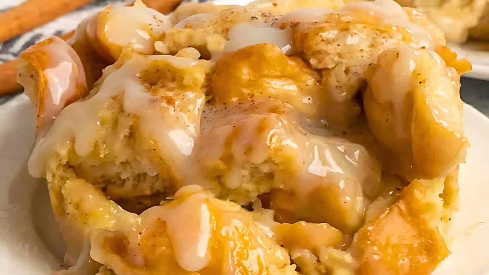 Close-up of a dessert featuring chunks of bread pudding drizzled with creamy glaze. The golden-brown pieces are moist and topped with a glossy, sweet sauce, adding an inviting shine to the dish. A cinnamon stick is partially visible in the background.