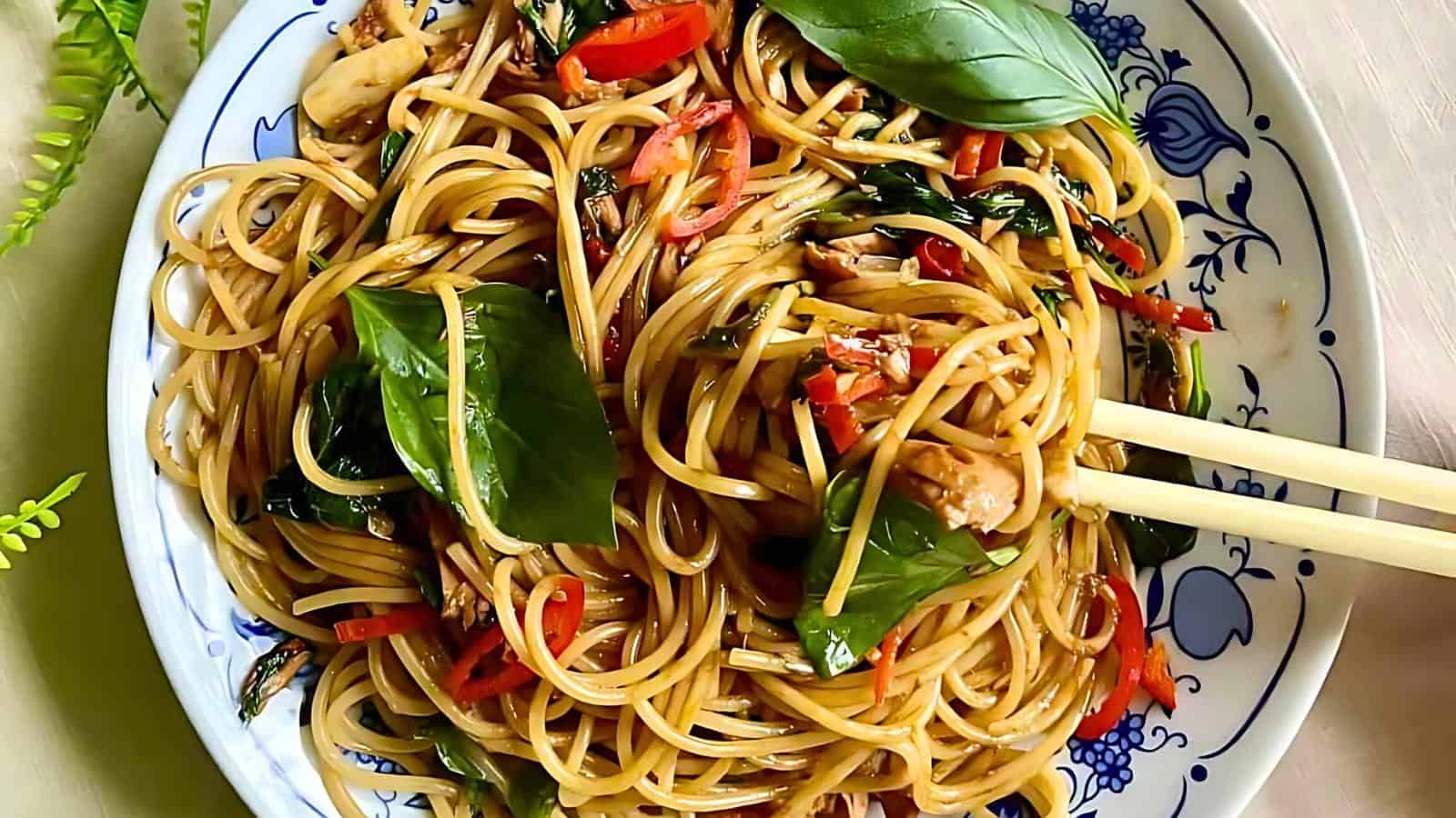 A close-up of a plate of stir-fried noodles with vegetables, garnished with fresh basil leaves. The noodles are intertwined with sliced red bell peppers, leafy greens, and other assorted vegetables, with chopsticks lifting a portion above the plate.