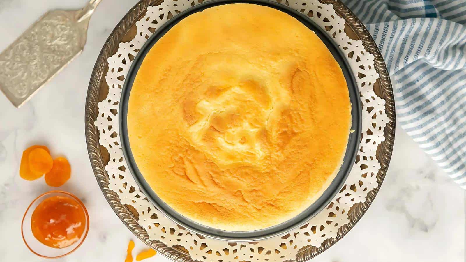 A round, golden-brown cheesecake sits on a decorative white lace-patterned tray. A pie server is on the left, and a small bowl of apricot glaze is nearby on the marble surface. A blue-striped cloth lies to the right.