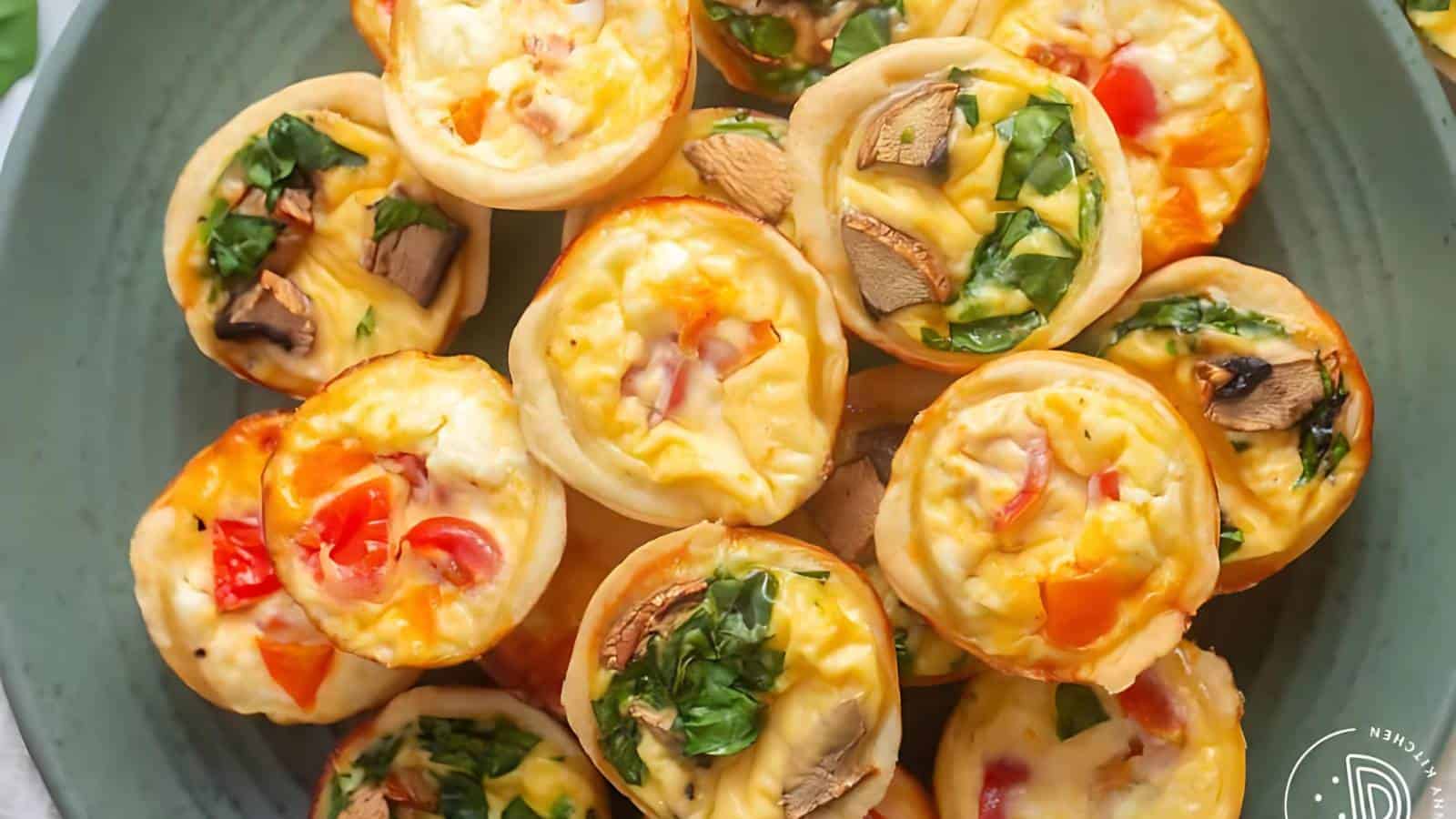 A plate of mini quiches topped with vegetables like tomatoes, mushrooms, and spinach, arranged attractively on a green ceramic platter. The quiches have golden, crisp edges and vibrant, colorful toppings.