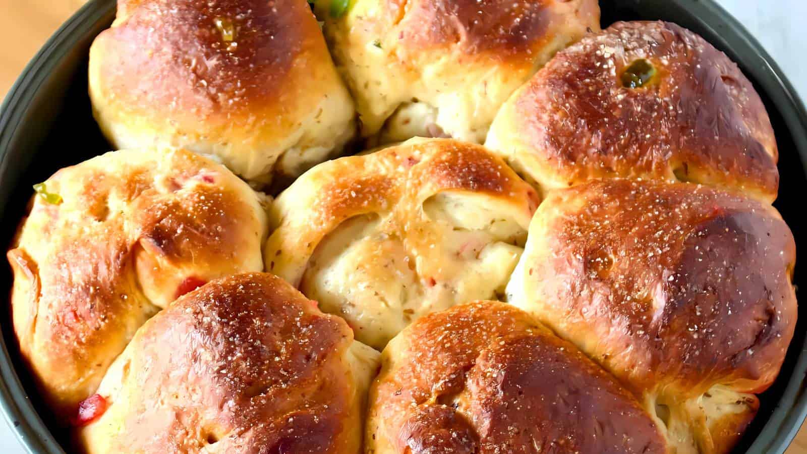 A close-up of freshly baked, golden-brown bread rolls in a round baking pan. The rolls have a shiny, slightly crusty exterior with sprinkles of herbs and salt visible on top.