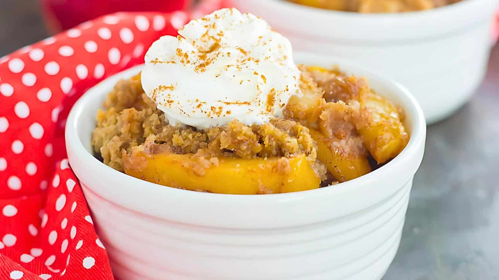 A small white ramekin filled with peach cobbler topped with a dollop of whipped cream and a sprinkle of cinnamon. A red polka-dotted napkin is beside the ramekin, adding a pop of color to the background.