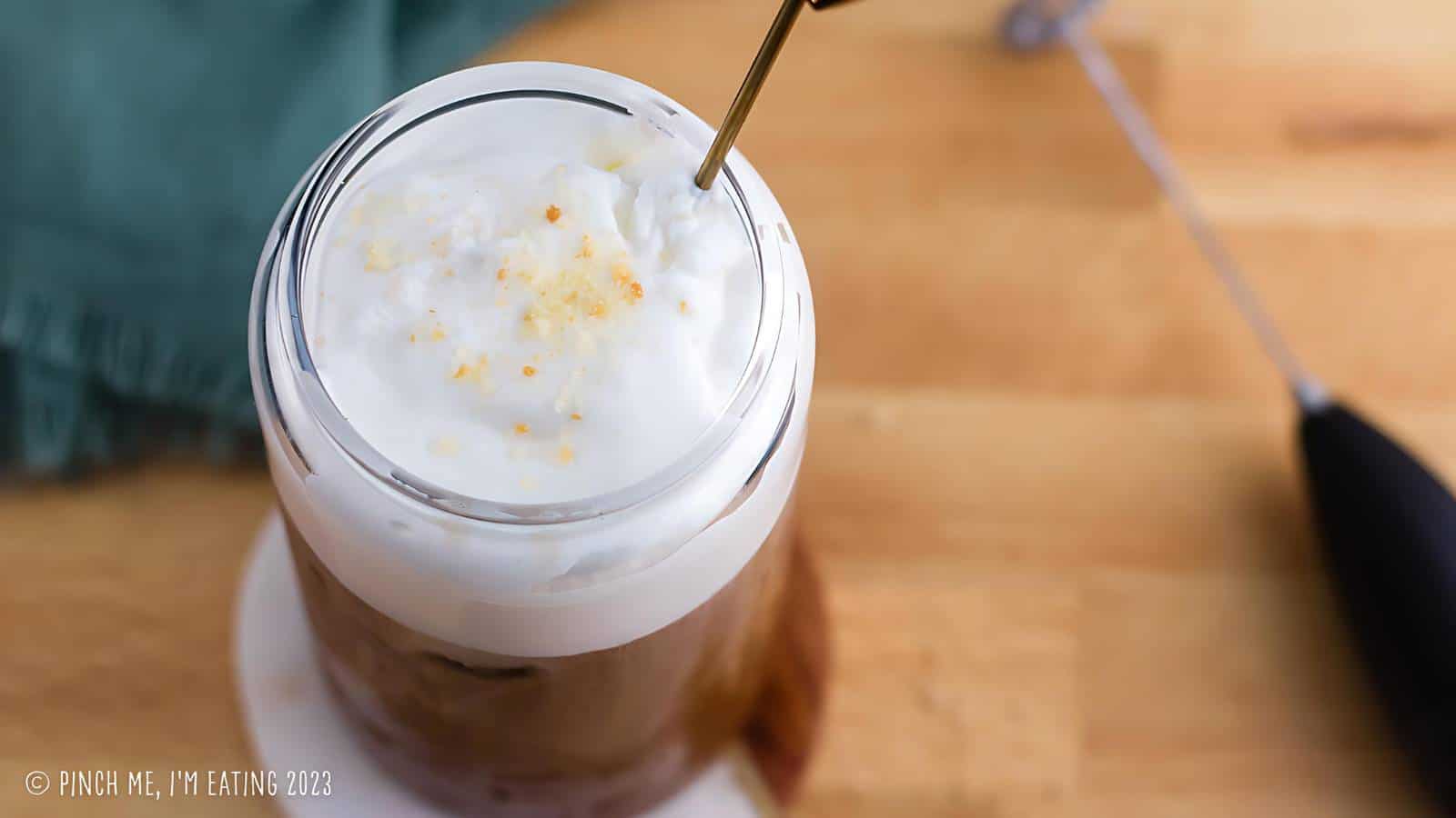 A glass filled with iced coffee topped with whipped cream is on a table next to a small whisk and green cloth.