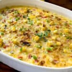 A baked casserole dish filled with a golden-brown mixture of diced vegetables, corn, and herbs, topped with fresh thyme and green onions, on a wooden surface.