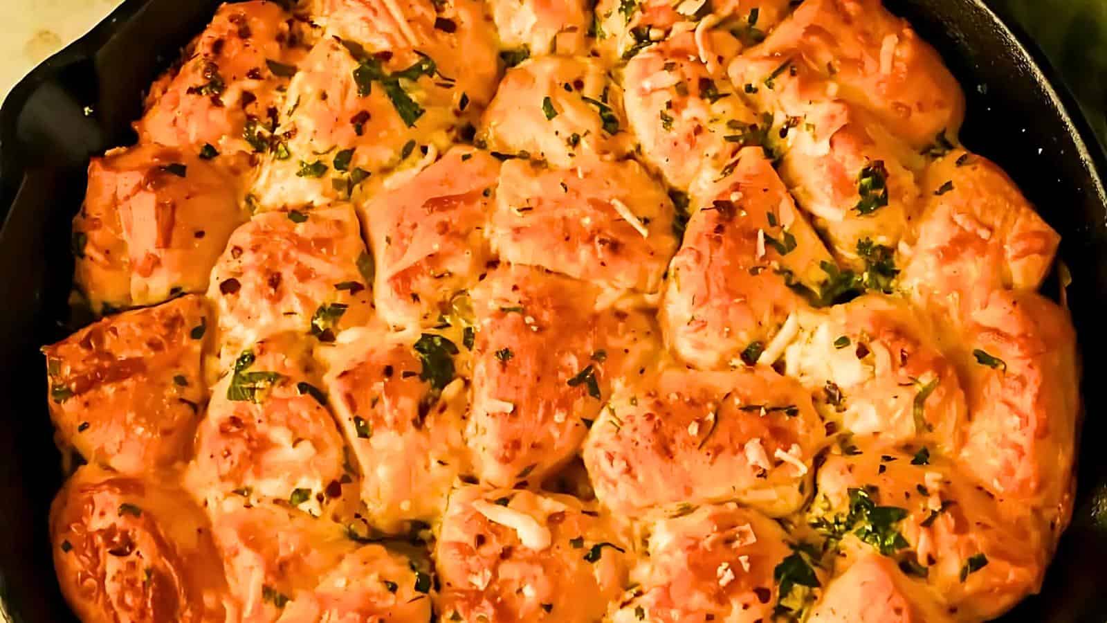 Baked pull-apart bread in a skillet, topped with melted cheese, bits of parsley, and red pepper flakes. The bread is golden brown and arranged closely together, creating a savory, appetizing dish.