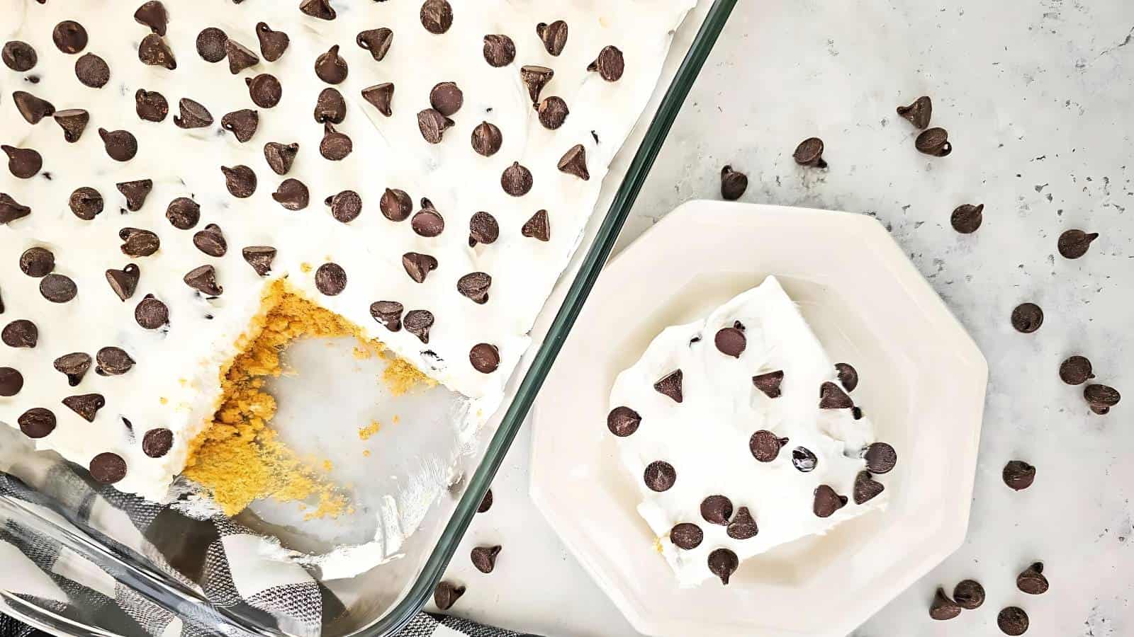 A tray of dessert topped with chocolate chips, featuring a partially eaten square piece on a plate. Exposed crumbly yellow layer underneath and scattered chocolate chips around.