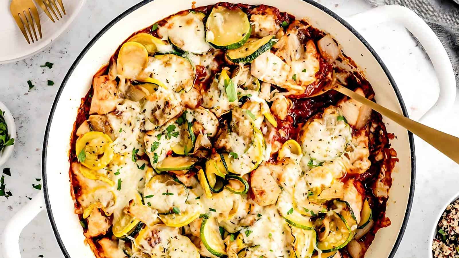 A pan of baked vegetable casserole topped with melted cheese, featuring sliced zucchini, yellow squash, and chopped herbs. A serving spoon is placed in the dish.