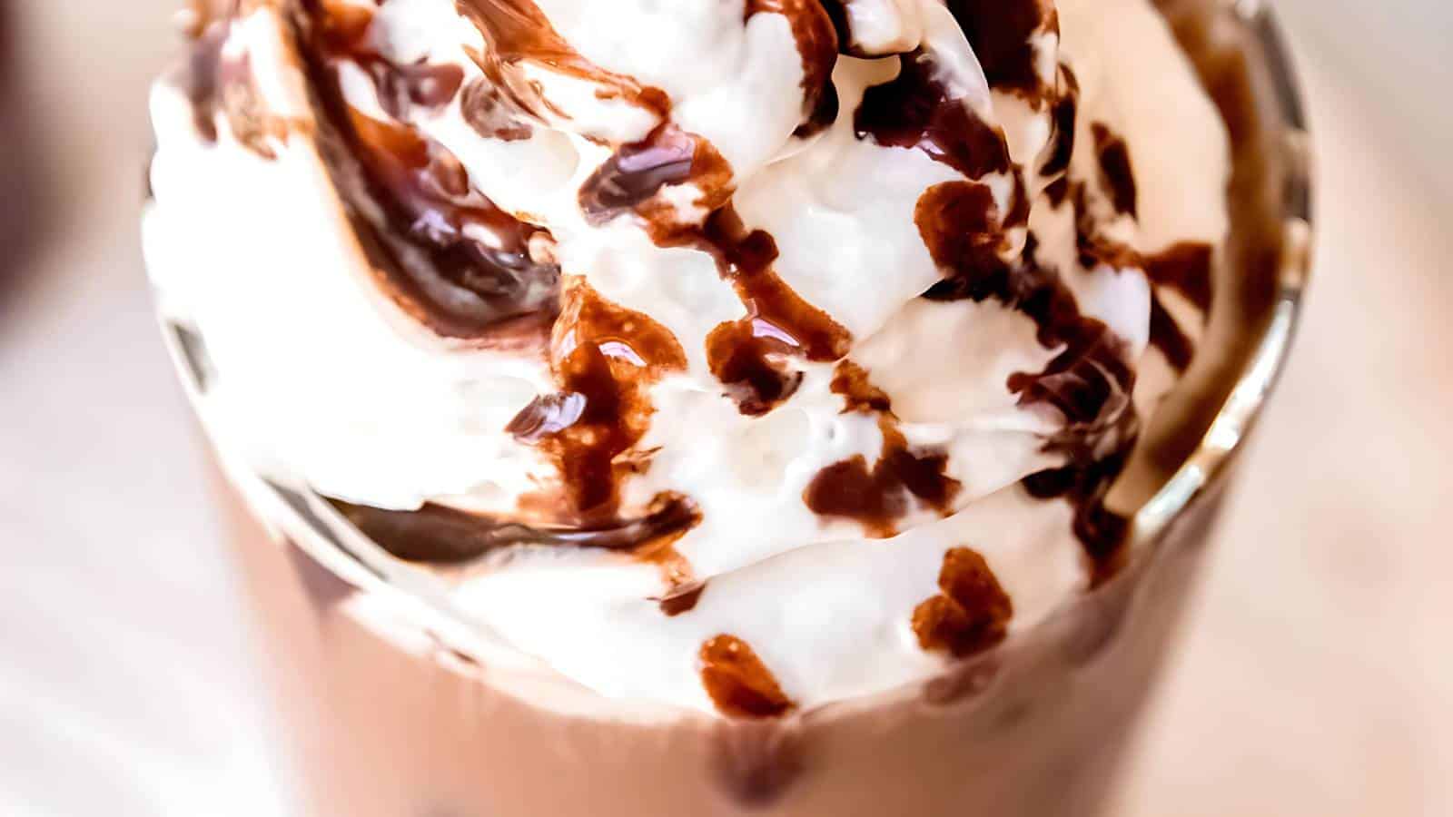 A glass filled with a creamy dessert, topped with whipped cream and drizzled with chocolate sauce.