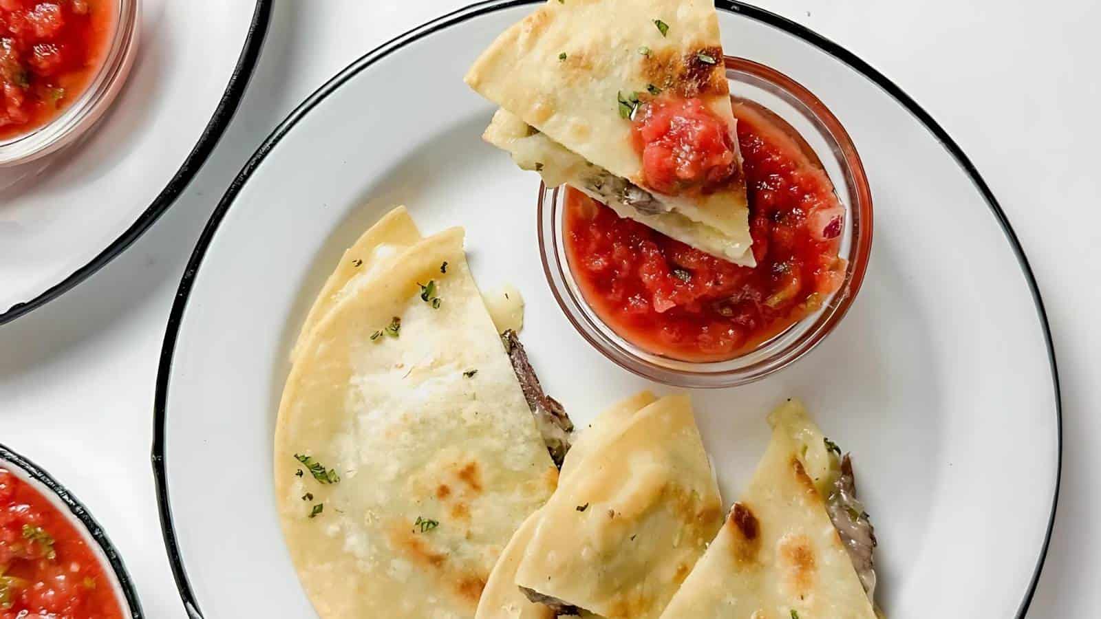 A plate of quesadillas filled with melted cheese and grilled meat, garnished with herbs. A small bowl of red salsa is served on the side. Several slices are positioned around the salsa, ready for dipping.