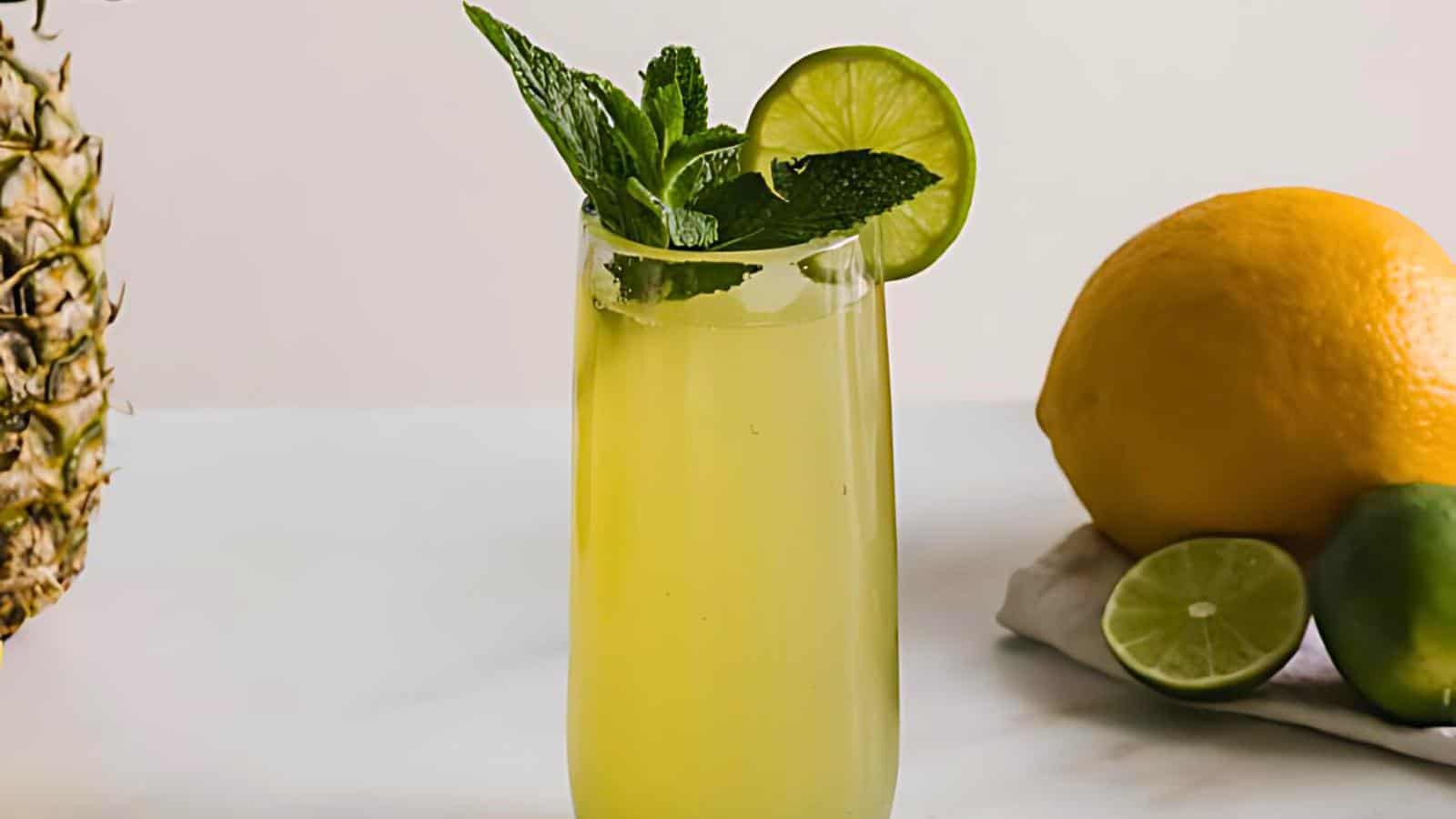 A tall glass of yellow beverage with a lime slice and mint leaves garnish, beside a pineapple, lemon, and sliced lime on a white surface.