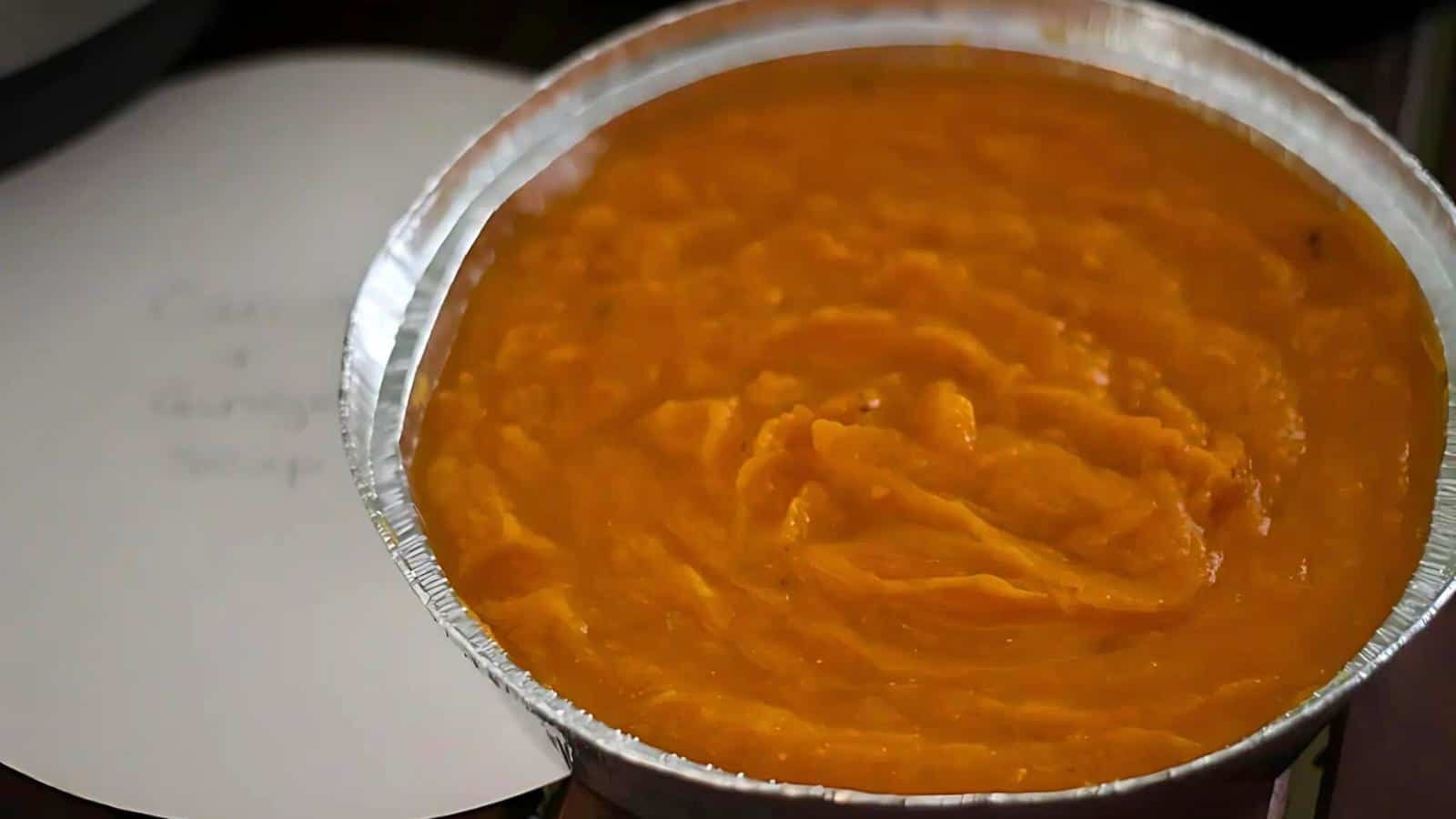 A foil container filled with orange-colored soup placed on a white round surface.