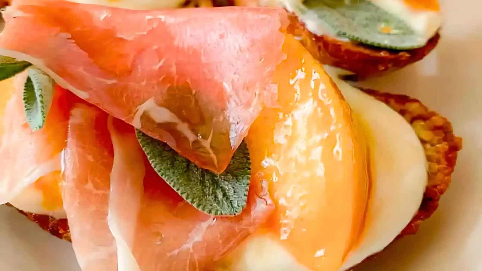 Cheese, sliced prosciutto, peach slices, and fresh herbs on a piece of bruschetta, served on a white plate.