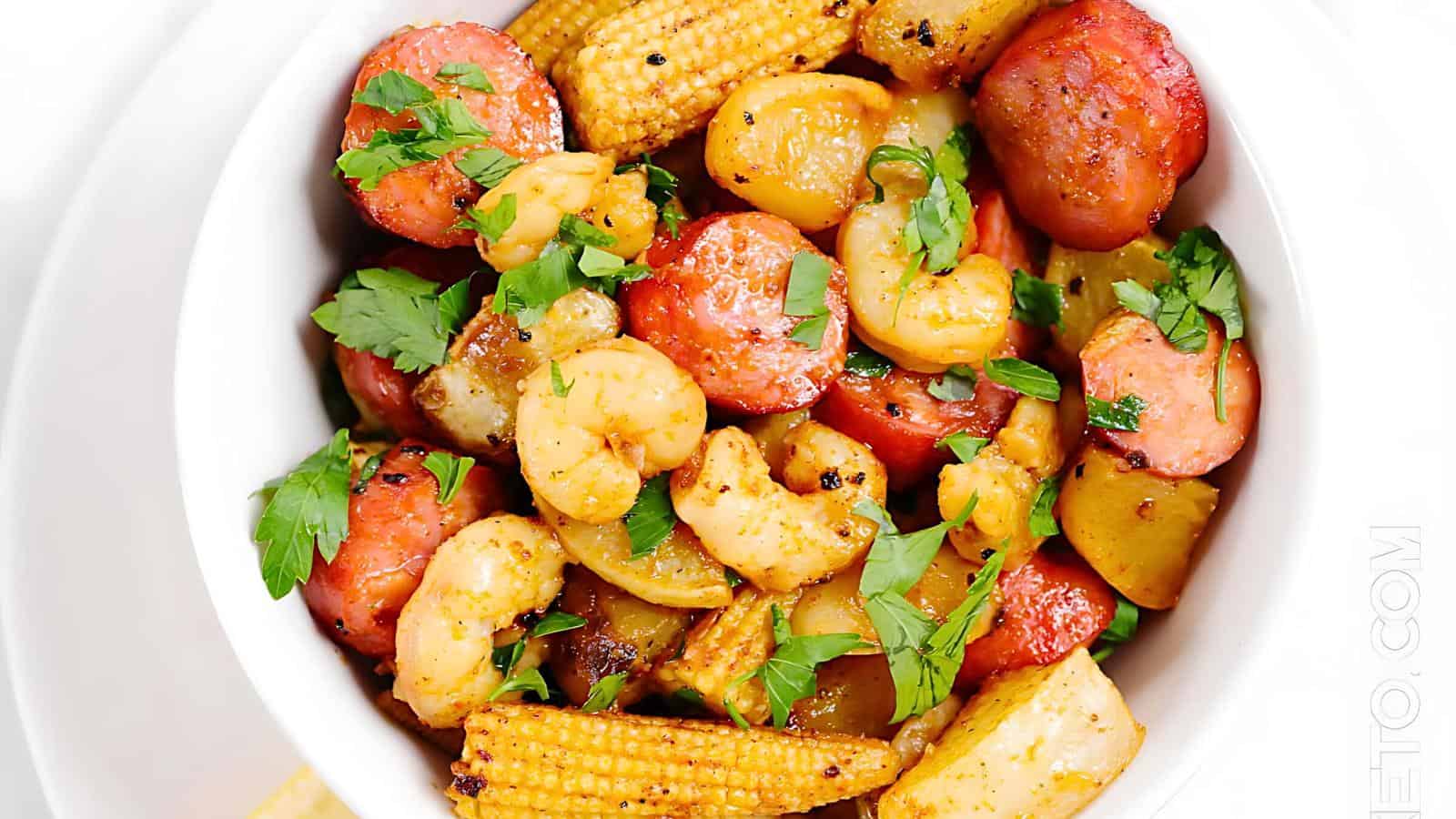 A bowl containing baby corn, sliced potatoes, shrimp, and sausage, garnished with chopped herbs.