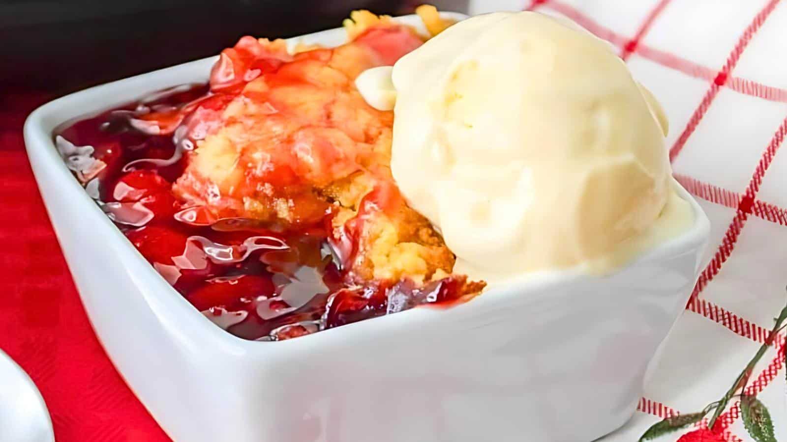 A close-up of a dessert in a white square dish featuring a crumble with a red berry sauce and a scoop of vanilla ice cream on top, set against a red and white checkered cloth background.