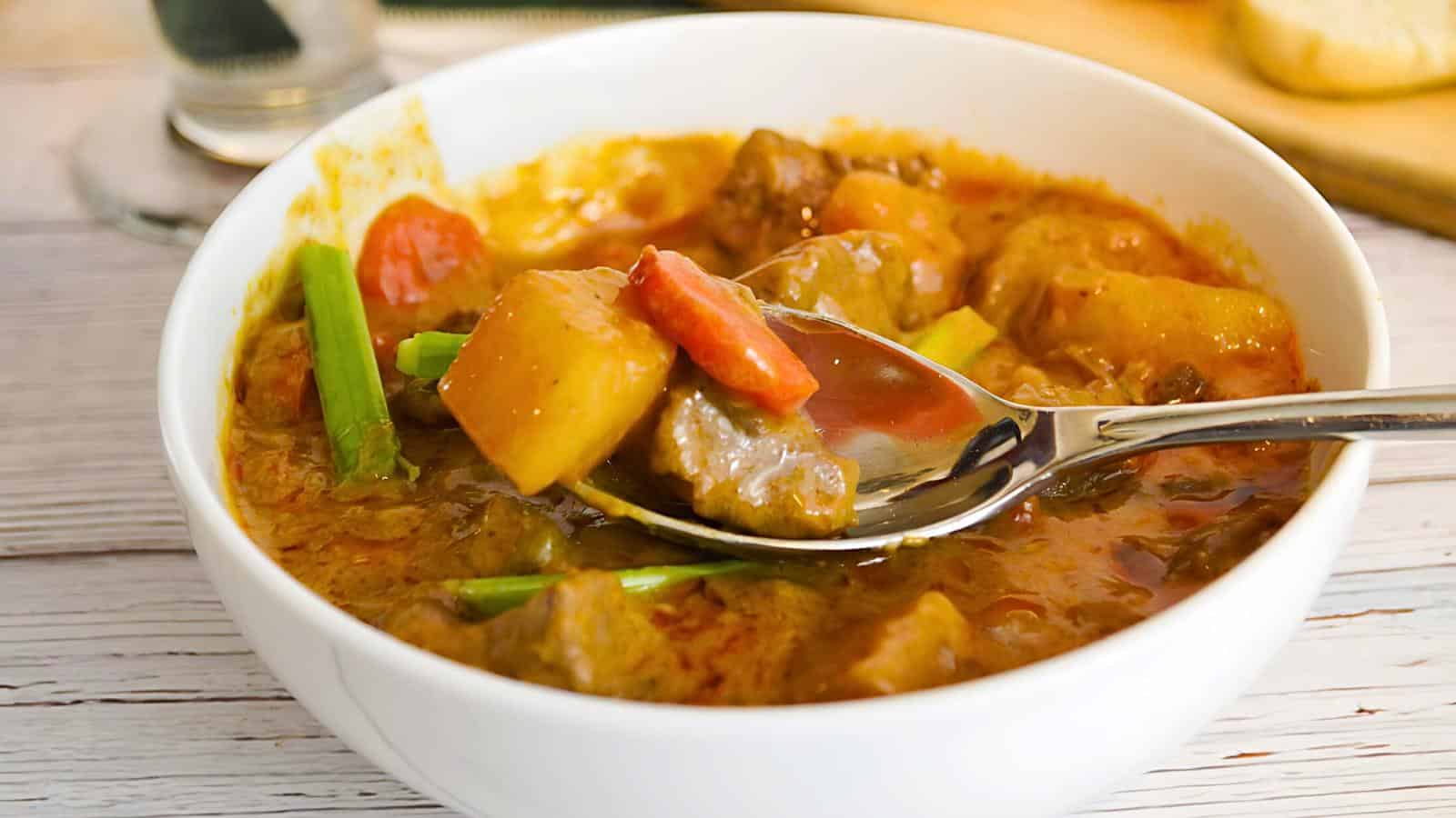 A white bowl filled with beef stew, including chunks of beef, carrots, potatoes, and green onions. A spoon in the bowl holds a portion of the stew.