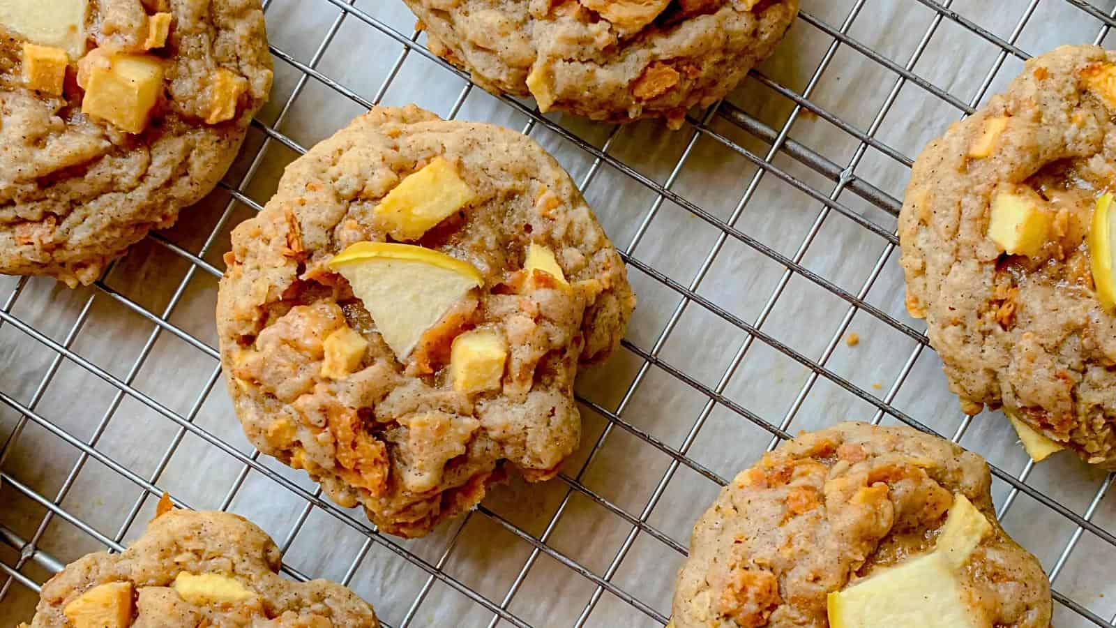 Close-up of several freshly baked apple cinnamon cookies cooling on a wire rack. The cookies are golden-brown with visible chunks of apple, and the rack sits on a light-colored surface. The cookies have a slightly crumbled texture, emphasizing their homemade look.