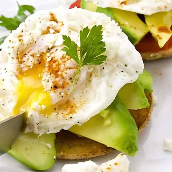 A poached egg with seasoning, sliced avocado, and parsley on a piece of toasted bread is being cut with a knife on a white plate. A lime wedge and another toast are in the background.