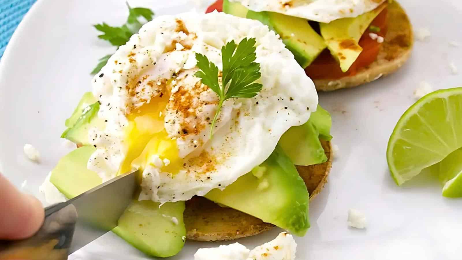 A poached egg with seasoning, sliced avocado, and parsley on a piece of toasted bread is being cut with a knife on a white plate. A lime wedge and another toast are in the background.