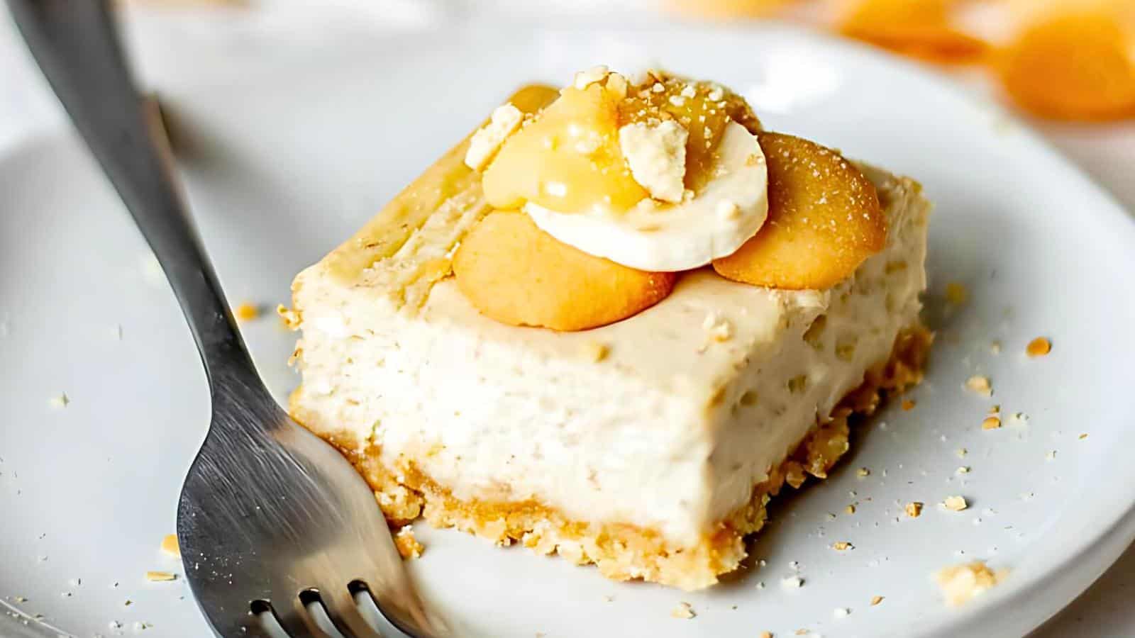 A slice of cheesecake topped with whipped cream, banana slices, and crumbled cookies, served on a white plate with a fork beside it.