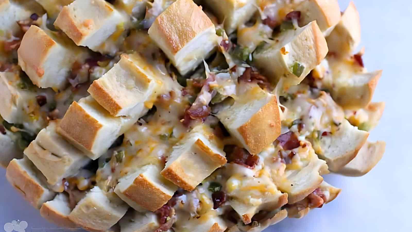 A baked loaf of bread has been scored in a crisscross pattern and stuffed with melted cheese, diced bacon, and green onions. The golden crust contrasts with the gooey filling, creating a savory pull-apart dish on a white background.