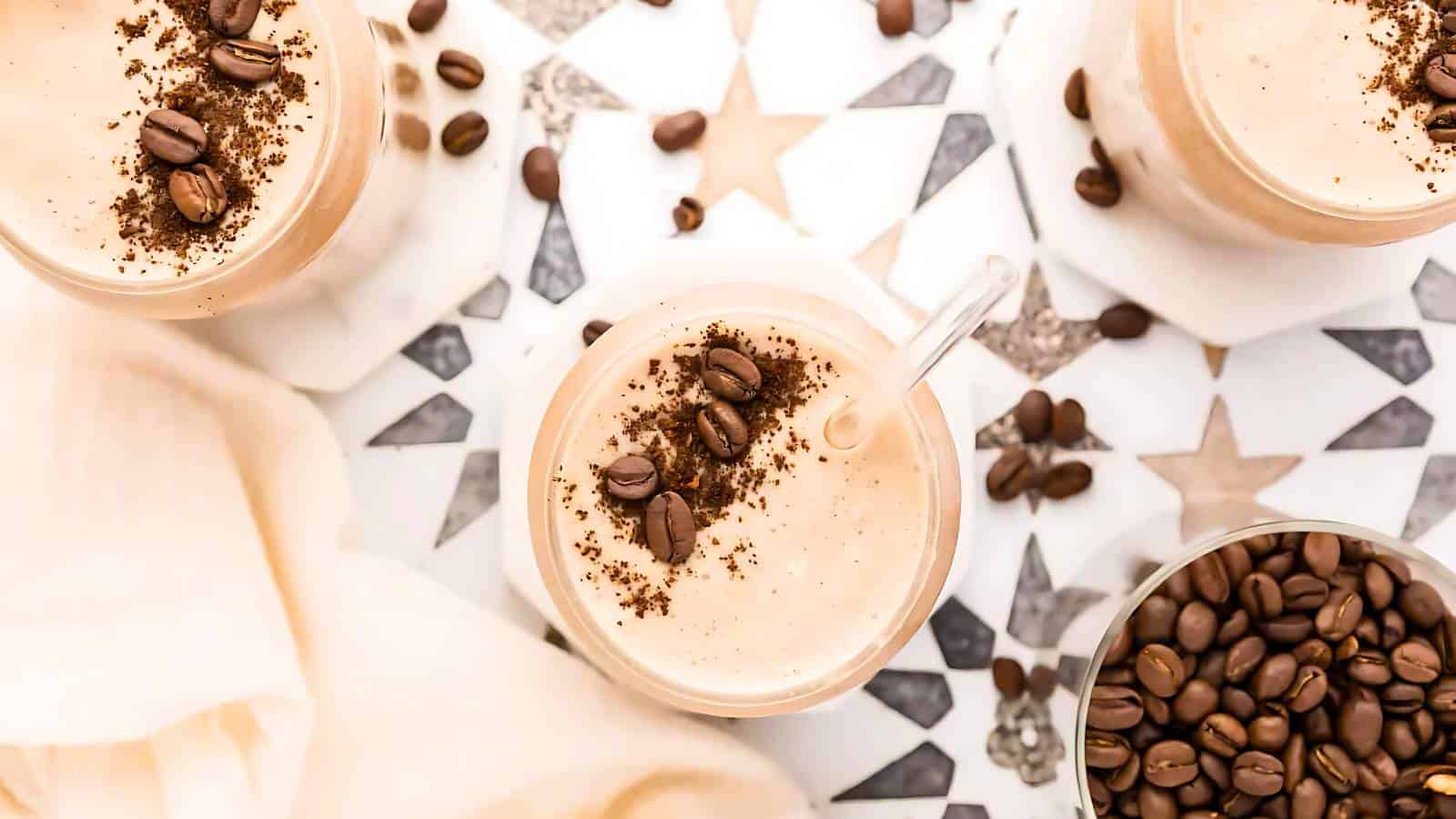 Three coffee-flavored milkshakes garnished with coffee beans and ground coffee. Bowl of coffee beans on the right, white cloth on the left, patterned surface in the background.