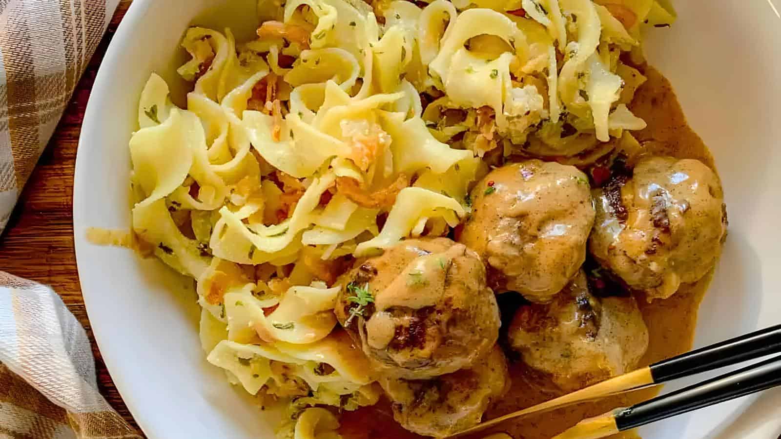 A white bowl holds creamy meatballs in brown sauce served over buttered egg noodles. A fork and knife are placed beside the bowl.