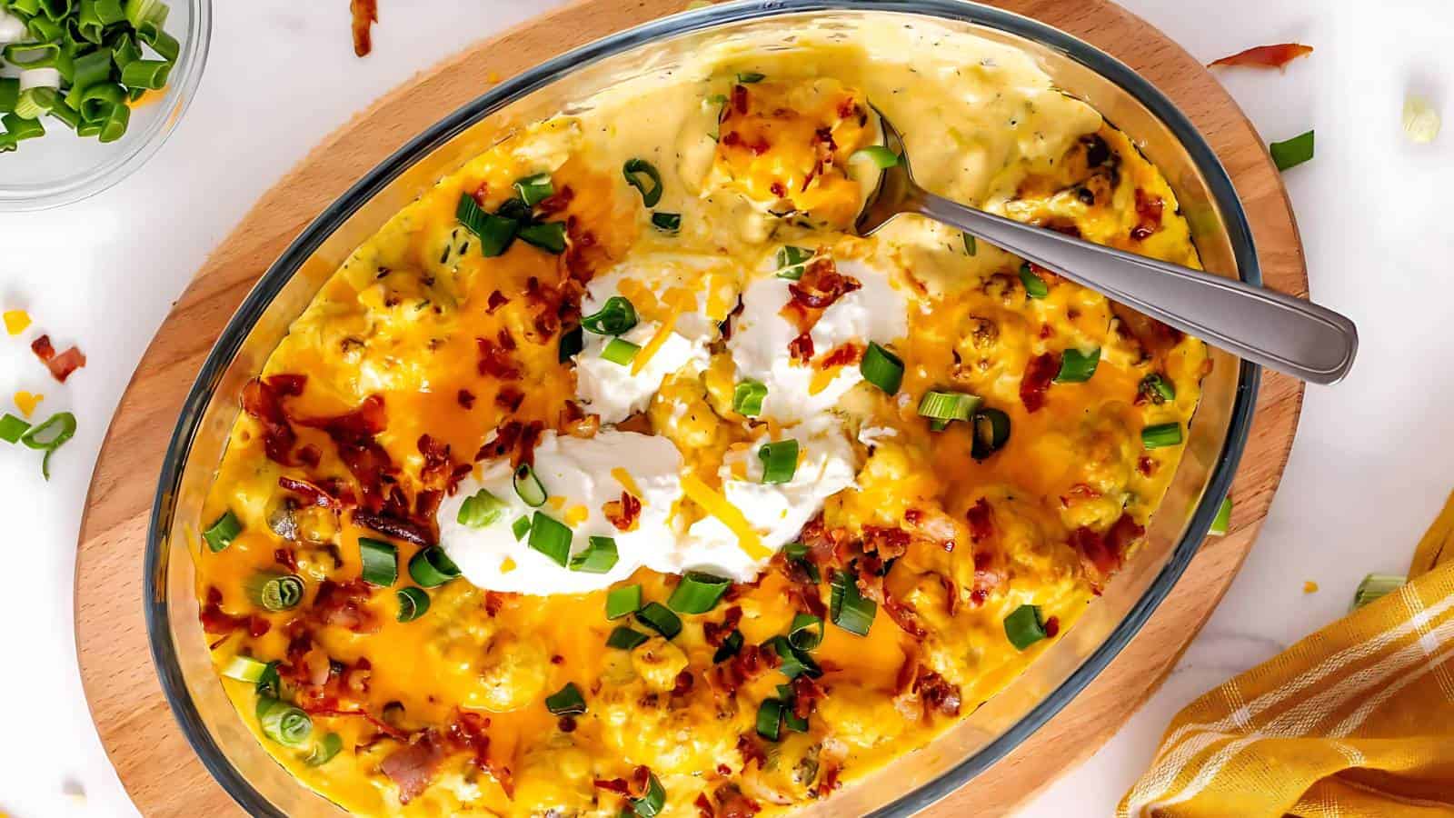 An oval dish of baked mashed potatoes topped with melted cheese, sour cream, chopped green onions, and bacon bits, with a serving spoon.