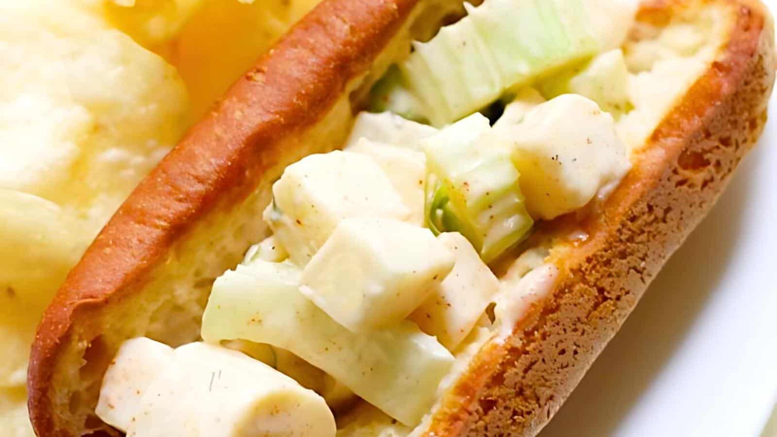 A sandwich roll filled with diced apples and celery in a creamy dressing, with potato chips partially visible in the background.