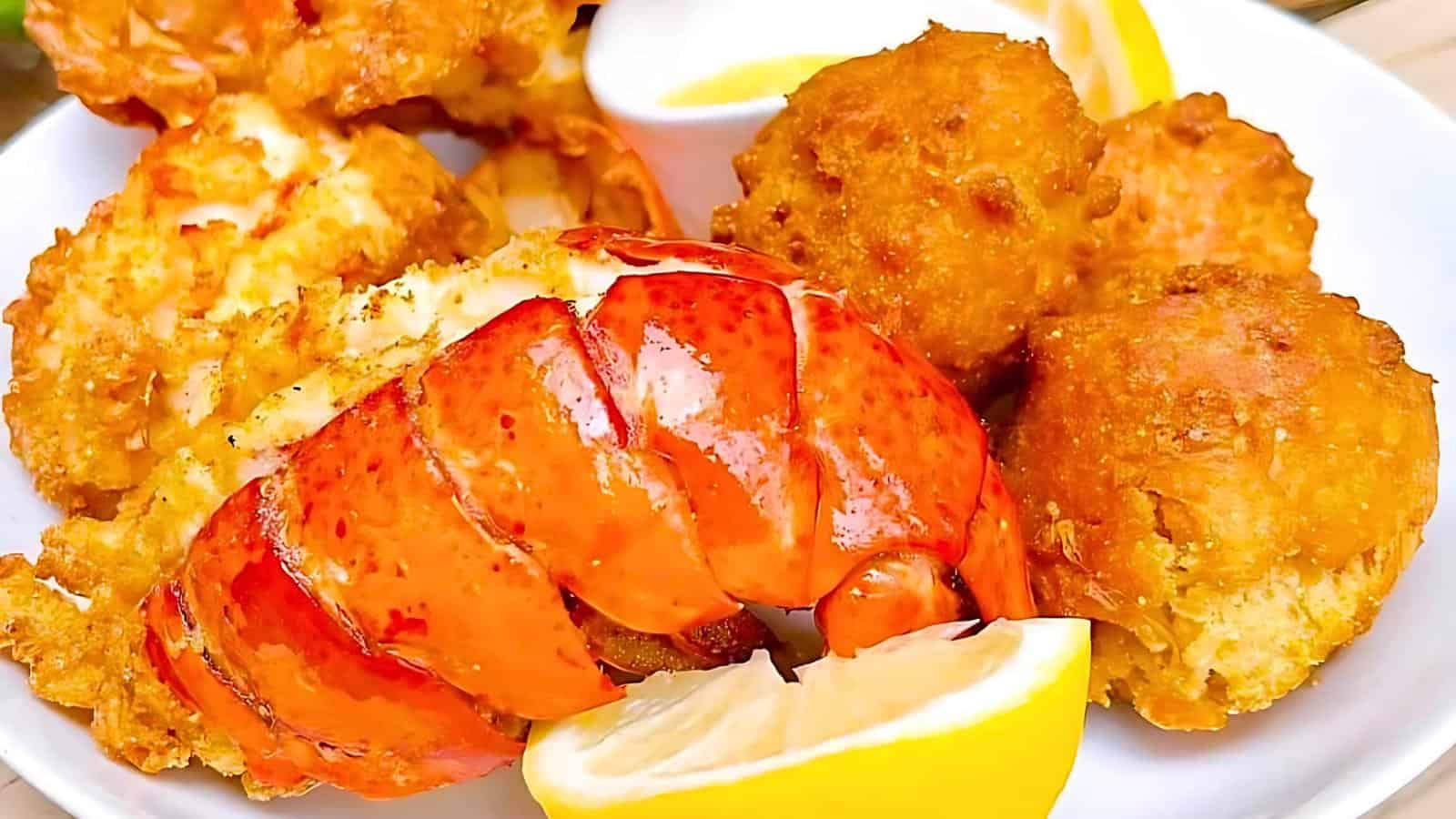 A white plate holds fried lobster tail, hush puppies, corn fritters, a lemon wedge, and a small cup of dipping sauce.