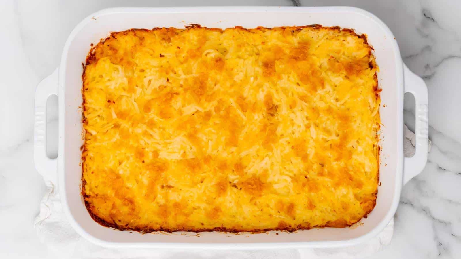 A baked potato casserole in a white rectangular dish, featuring a golden-brown, cheesy top layer. The dish rests on a marble surface.