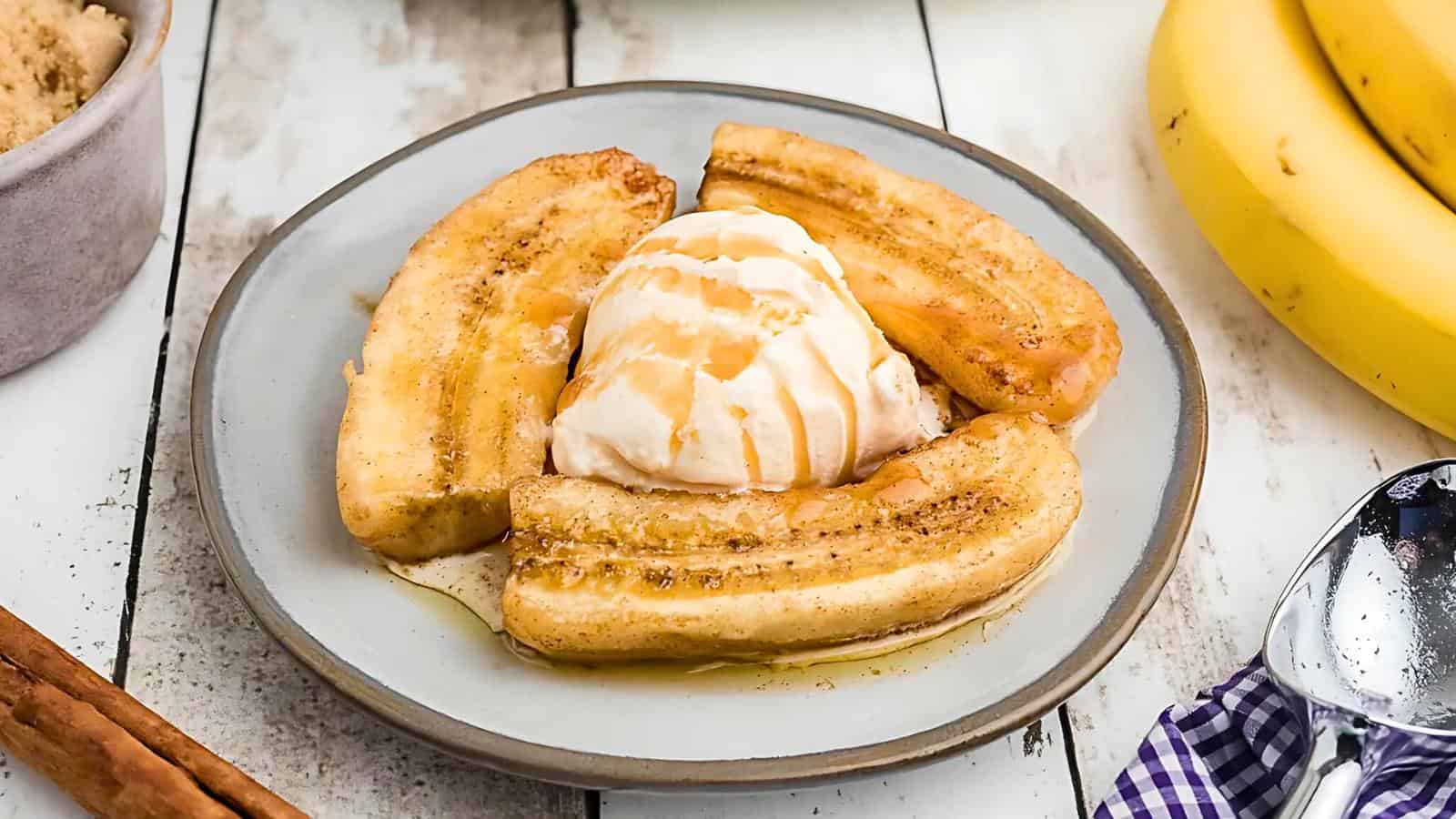 A plate of caramelized banana slices topped with a scoop of vanilla ice cream and drizzled with caramel sauce. The dessert is set on a rustic wooden table, with whole bananas, brown sugar, and a cinnamon stick nearby.