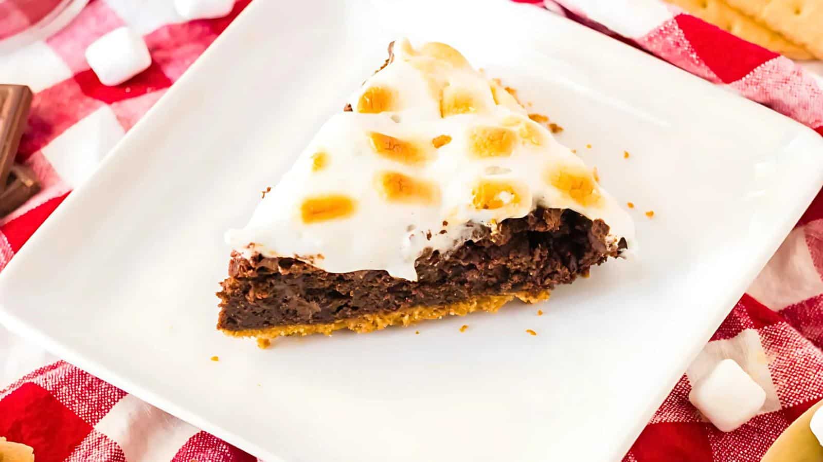 A slice of chocolate dessert with a layer of toasted marshmallows on top, served on a square white plate on a red and white checkered tablecloth.